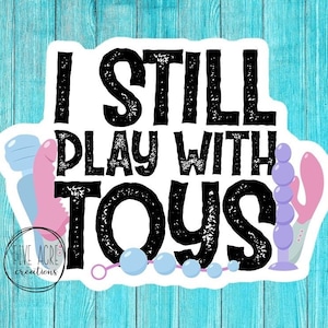 May include: A white sticker with black text that reads "I still play with toys". The sticker features a pink and blue toy with a purple beaded tail.