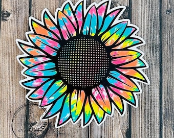 May include: A tie-dye sunflower with pink, blue, yellow, and orange colors. The sunflower has a black center with a white outline.