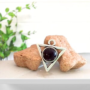 May include: Silver-toned pendant with a triangular frame and a dark, round center stone. The pendant is displayed on a light-colored surface with two rough, tan-colored stones in the background. The pendant has a small loop at the top for a chain.