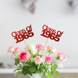May include: Two red glitter 'Class of 1965' signs are stuck into a bouquet of pink and white roses in a white vase.