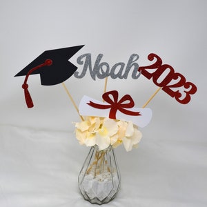 Puede incluir: Centro de mesa de graduaci&oacute;n con un gorro de graduaci&oacute;n negro, un diploma blanco con una cinta roja y letras de purpurina plateadas que deletrean "Noah 2023" en palos de madera en un jarr&oacute;n de vidrio transparente con flores blancas.
