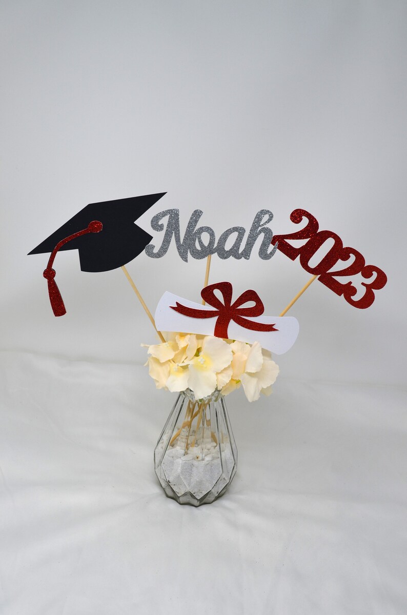 Puede incluir: Centro de mesa de graduaci&oacute;n con un gorro de graduaci&oacute;n negro, un diploma blanco con una cinta roja y letras de purpurina plateadas que deletrean "Noah 2023" en palos de madera en un jarr&oacute;n de vidrio transparente con flores blancas.