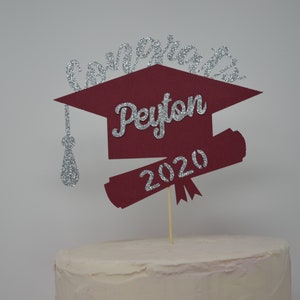Puede incluir: Topper de pastel de graduaci&oacute;n de color burdeos en forma de gorro y diploma con detalles de purpurina plateada. El topper dice "Congrats Peyton 2020".
