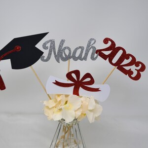 Puede incluir: Centro de mesa de graduaci&oacute;n con un gorro de graduaci&oacute;n negro, un letrero de "Noah 2023" con purpurina plateada, un diploma blanco con un lazo rojo y flores blancas en un jarr&oacute;n transparente.