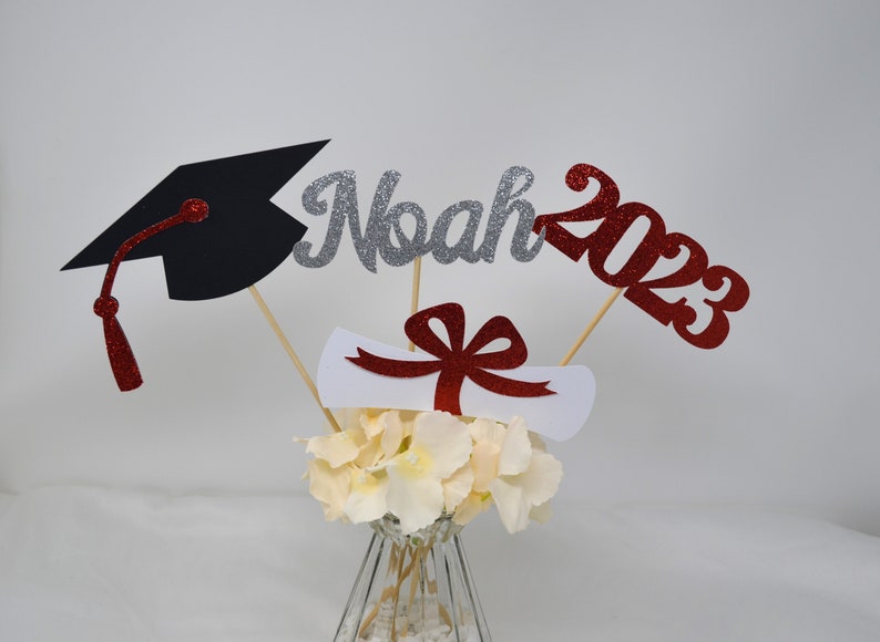 Puede incluir: Centro de mesa de graduaci&oacute;n con un gorro de graduaci&oacute;n negro, un letrero de "Noah 2023" con purpurina plateada, un diploma blanco con un lazo rojo y flores blancas en un jarr&oacute;n transparente.