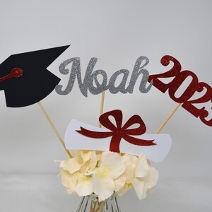 Puede incluir: Centro de mesa de graduaci&oacute;n con un gorro de graduaci&oacute;n negro, un nombre "Noah" de purpurina plateada, un "2023" de purpurina roja, un diploma blanco con una cinta roja y un ramo de flores blancas.