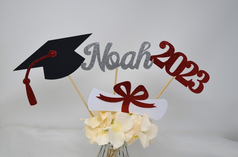 Puede incluir: Centro de mesa de graduaci&oacute;n con un gorro de graduaci&oacute;n negro, un nombre "Noah" de purpurina plateada, un "2023" de purpurina roja, un diploma blanco con una cinta roja y un ramo de flores blancas.