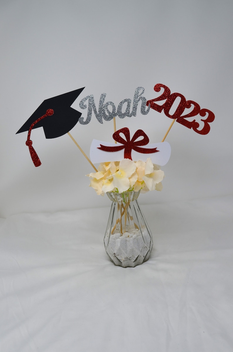 Puede incluir: Centro de mesa de graduaci&oacute;n con un gorro de graduaci&oacute;n negro, un diploma blanco con una cinta roja, un "Noah" de purpurina plateada y un "2023" de purpurina roja en palos de madera en un jarr&oacute;n de vidrio transparente con flores blancas.