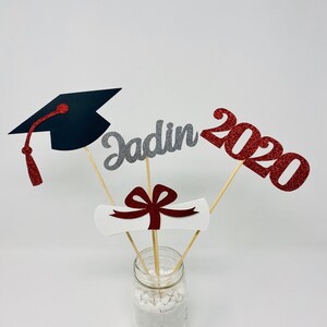 Puede incluir: Centro de mesa de graduaci&oacute;n con un gorro de graduaci&oacute;n negro, un diploma blanco con un lazo rojo, un "Jadin" de purpurina plateada y un "2020" de purpurina roja en un frasco de vidrio transparente con arena blanca.