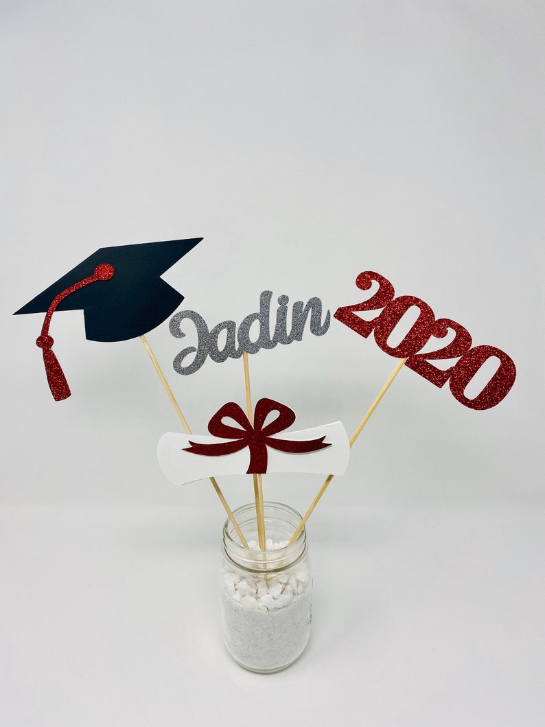 Puede incluir: Centro de mesa de graduaci&oacute;n con un gorro de graduaci&oacute;n negro, un diploma blanco con un lazo rojo, un "Jadin" de purpurina plateada y un "2020" de purpurina roja en un frasco de vidrio transparente con arena blanca.