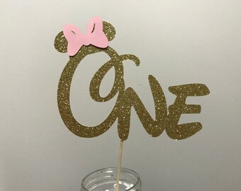 May include: Gold glitter "one" cake topper with a pink bow on top. The topper is on a wooden stick and is perfect for a first birthday party.