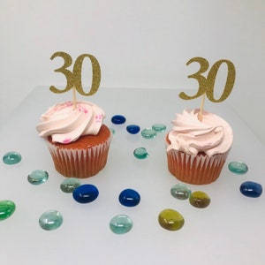 Puede incluir: Dos cupcakes con glaseado rosa y adornos de cupcake de purpurina dorada "30". Los cupcakes están sobre una superficie blanca con pequeñas piedras de vidrio de colores.
