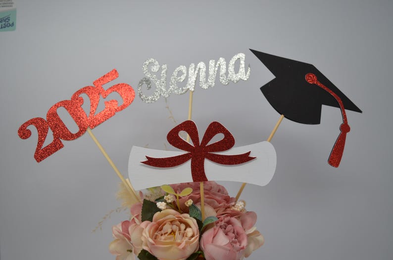 Puede incluir: Centro de mesa de graduaci&oacute;n con 2025 en purpurina roja, Sienna en purpurina plateada, un lazo de purpurina roja en un diploma blanco y un gorro de graduaci&oacute;n negro con borla roja.