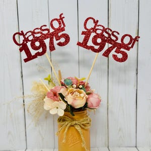 May include: Two red glitter Class of 1975 signs in a mason jar with pink and white flowers.