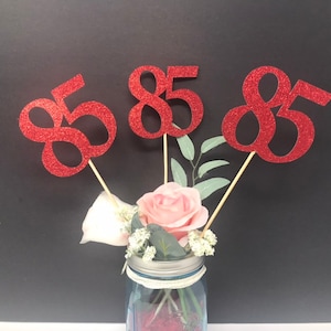 May include: Three red glitter number 85s on sticks in a mason jar with flowers.