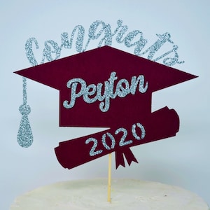 Puede incluir: Un adorno para pastel de gorro de graduaci&oacute;n burdeos con un borla de purpurina plateada y las palabras "Congrats Peyton 2020" en purpurina plateada.