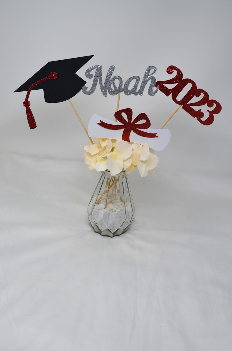 Puede incluir: Centro de mesa de graduaci&oacute;n con un gorro de graduaci&oacute;n negro, un diploma blanco con una cinta roja, un 'Noah' de purpurina plateada y un '2023' de purpurina roja en palos de madera en un jarr&oacute;n de vidrio transparente con flores blancas.