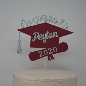 Puede incluir: Un adorno para pastel de graduaci&oacute;n de color burdeos en forma de gorro y diploma con letras plateadas brillantes. El adorno dice "Congrats Peyton 2020".