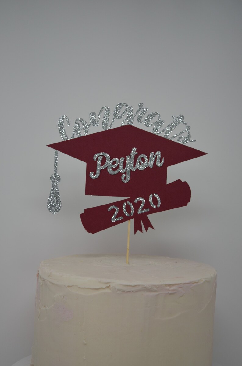 Puede incluir: Un adorno para pastel de graduaci&oacute;n de color burdeos en forma de gorro y diploma con letras plateadas brillantes. El adorno dice "Congrats Peyton 2020".