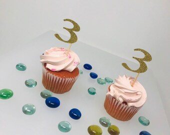 May include: Two pink cupcakes with white frosting and gold glitter number 3 cupcake toppers.