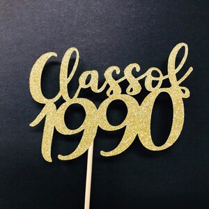 Puede incluir: Topper para pastel de purpurina dorada con el texto "Class of 1990" en una fuente cursiva.