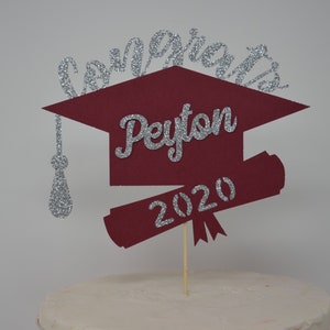 Puede incluir: Topper para pastel de graduaci&oacute;n en forma de gorro de color burdeos con letras de purpurina plateada. El topper dice "Congrats Peyton 2020".