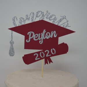 Puede incluir: Un adorno para pastel de graduaci&oacute;n rojo y plateado con un gorro de graduaci&oacute;n, un diploma y el texto "Congrats Peyton 2020".