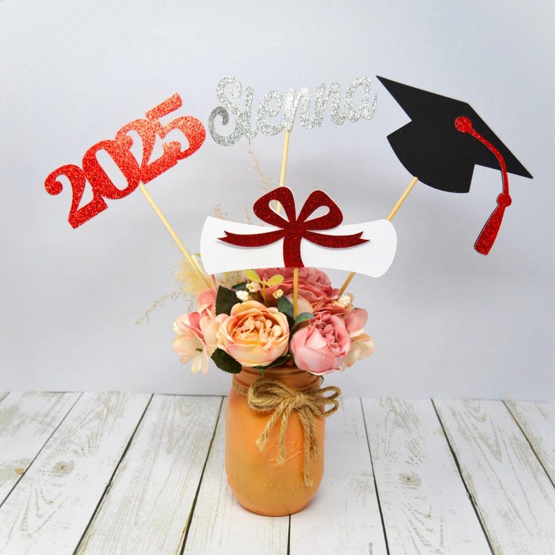 Puede incluir: Un centro de mesa de graduaci&oacute;n con un jarr&oacute;n de tarro de alba&ntilde;il lleno de flores rosas y melocot&oacute;n. El centro de mesa incluye un 2025 de purpurina roja, un "Sierra" de purpurina plateada, un diploma blanco con un lazo rojo y un gorro de graduaci&oacute;n negro.