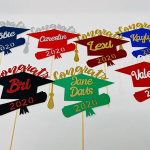 Puede incluir: Seis adornos para gorros de graduaci&oacute;n con los nombres Abbie, Carestin, Lexi, Kaylyn, Brl y Jane Davis, y el a&ntilde;o 2020. Los adornos est&aacute;n hechos de cartulina con purpurina en varios colores, incluyendo rojo, azul, verde y negro.