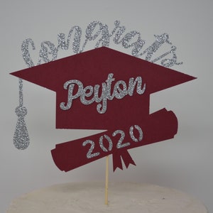 Puede incluir: Topper de pastel de graduaci&oacute;n de color burdeos con un acabado de purpurina plateada. El topper presenta el nombre "Peyton" y el a&ntilde;o "2020" en purpurina plateada.