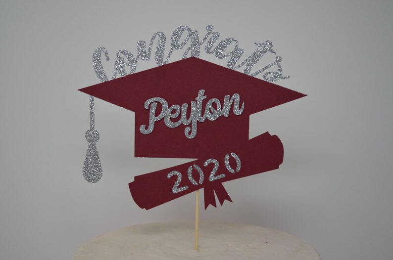 Puede incluir: Topper de pastel de graduaci&oacute;n de color burdeos con un acabado de purpurina plateada. El topper presenta el nombre "Peyton" y el a&ntilde;o "2020" en purpurina plateada.