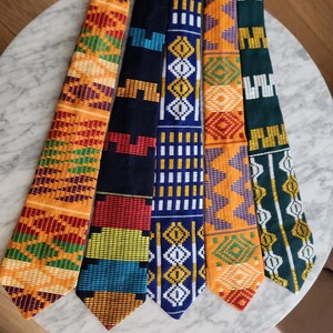May include: Five patterned neckties in a variety of colours, including orange, blue, green, and red. Each tie displays a unique geometric design, blending traditional and modern styles. The ties are arranged on a marble surface, showcasing their intricate patterns.