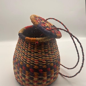 Op de afbeelding: Een geweven mand met een deksel, met een mix van rode, oranje, paarse en bruine kleuren. De mand heeft een ronde vorm met een deksel dat gedeeltelijk open is, waardoor de binnenkant zichtbaar is. Een gevlochten koordhandvat is aan het deksel bevestigd.