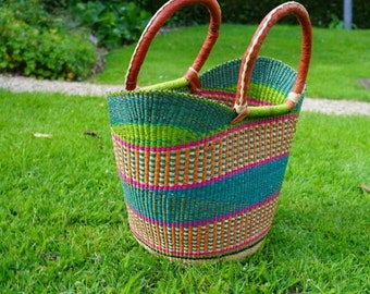 Pode incluir: Uma cesta tecida colorida com alças de couro. A cesta é verde, laranja, rosa e azul. Está sentada em uma superfície de grama verde.