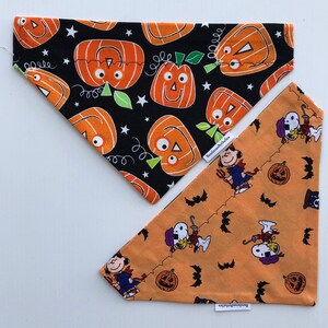 May include: Two reversible dog bandanas. One side is black with orange pumpkins and stars. The other side is orange with a pattern of Peanuts characters and pumpkins. The bandanas are labeled "Handmade by the Sea".