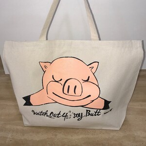 May include: Off-white canvas tote bag with a cartoon pig illustration. The pig is pink with black outlines and the text "Watch Out 4 my Butt" is printed below. The bag has two beige handles.