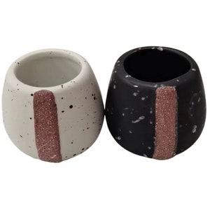 Może przedstawiać: Dwa małe, okrągłe doniczki ceramiczne z chropowatym wykończeniem. Jedna doniczka jest biała z czarnymi plamkami, a druga jest czarna z białymi plamkami. Obie doniczki mają pionowy różowy pasek.