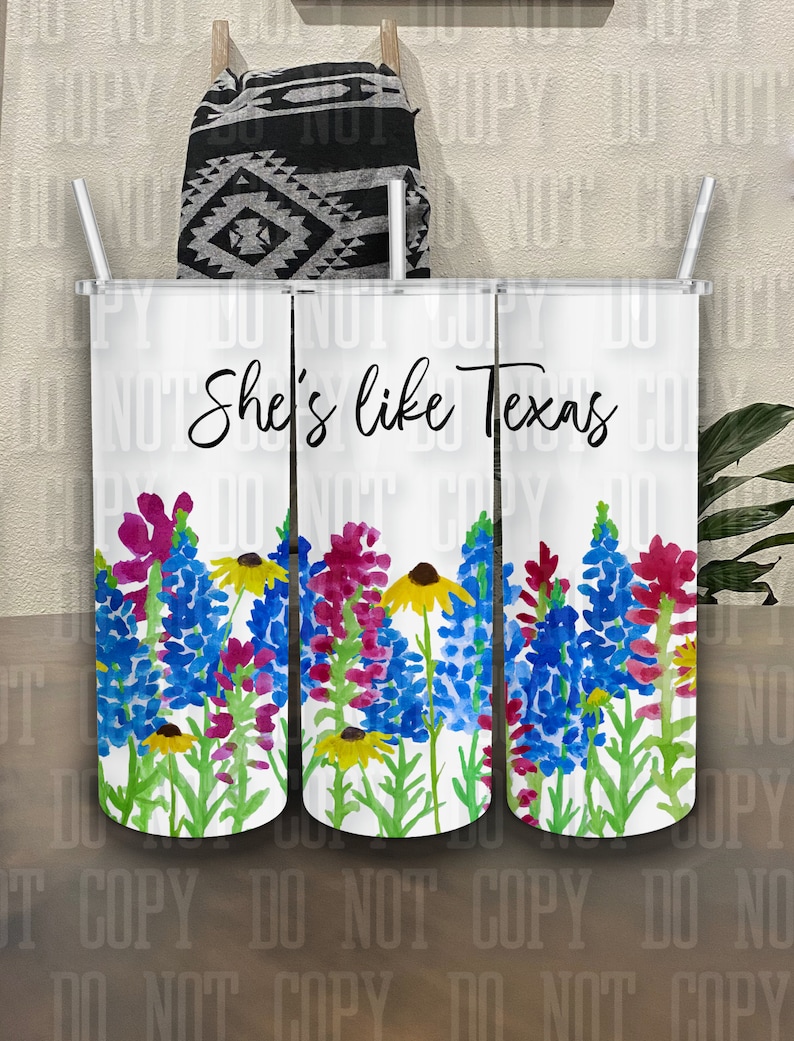 May include: Three white tumblers with a floral design and the text "She's like Texas". The flowers are blue, pink, yellow, and green. The tumblers have white lids and clear straws.