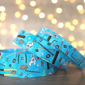 May include: Three blue fabric belts with a repeating pattern of tools, including wrenches, hammers, and screws. The belts are layered on top of each other, with a blurred background of golden bokeh lights.