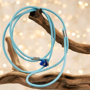 May include: A light blue necklace with a circular design, featuring silver-colored metal accents. The necklace is displayed on a piece of driftwood against a blurred background of soft, warm lights. The necklace has a small blue pendant.