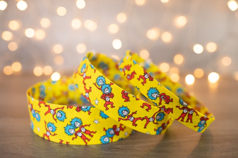 May include: Three yellow headbands with a repeating pattern of the characters from the book "The Cat in the Hat" by Dr. Seuss. The characters are wearing red and blue outfits and are dancing on a yellow background.