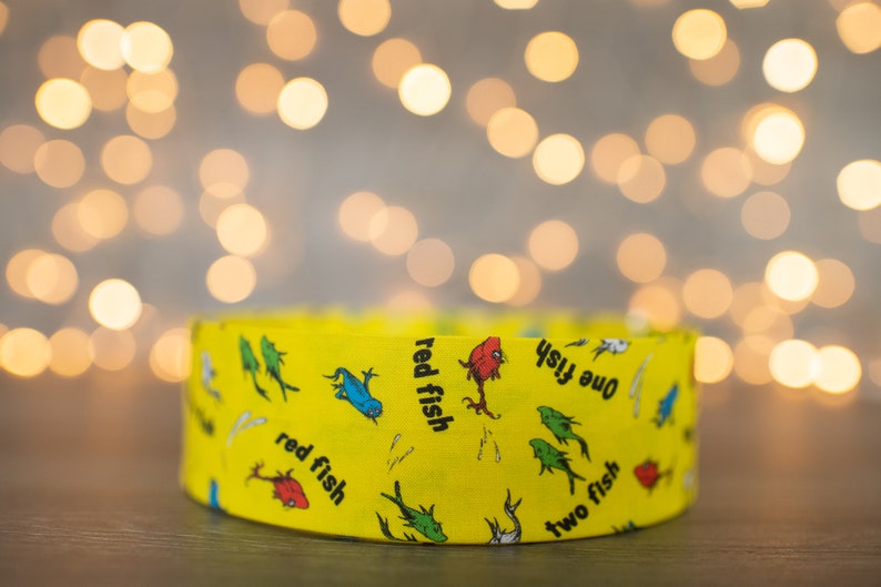 May include: A yellow fabric headband with a repeating pattern of red fish, blue fish, and green fish. The text "one fish, two fish, red fish, blue fish" is printed on the headband.