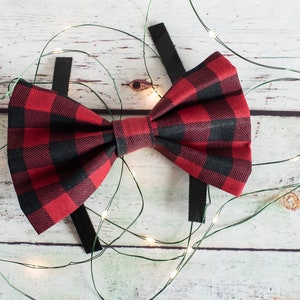 May include: A red and black plaid bow tie with black adjustable straps. The bow tie is on a white wooden background with green string lights.