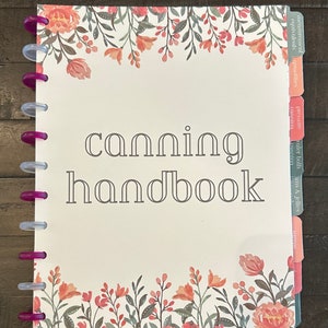 Puede incluir: Un manual blanco con el título "canning handbook" en texto gris. La portada tiene un borde floral con flores rosas y naranjas. El libro tiene una encuadernación de plástico transparente con anillos rosas. El borde derecho del libro tiene pestañas con las siguientes etiquetas: "agitación", "mermeladas y jaleas", "baño maría", "conservación", "presión", "cantidades", "equivalentes", "medición".