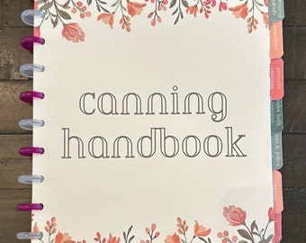 Puede incluir: Un manual blanco con el título "canning handbook" en texto gris. La portada tiene un borde floral con flores rosas y naranjas. El libro tiene una encuadernación de plástico transparente con anillos rosas. El borde derecho del libro tiene pestañas con las siguientes etiquetas: "agitación", "mermeladas y jaleas", "baño maría", "conservación", "presión", "cantidades", "equivalentes", "medición".