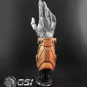 May include: A metallic, articulated robotic hand sculpture with a bronze-colored forearm and a black base. The hand is silver and detailed, with visible mechanical joints. The forearm has the text "GSI".