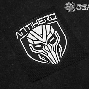 May include: Black patch with the word "ANTIHERO" in white block letters above a stylized white graphic of a mask. The patch is square with rounded corners and is set against a black background.