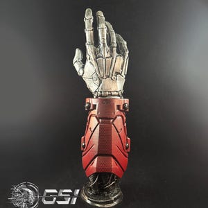 May include: A metallic silver and red robotic arm with a detailed skeletal hand design. The arm features a red segmented forearm and a circular base. The text "* MANNEQUIN HAND NOT INCLUDED" is visible at the top. The logo "GSI" is at the bottom.