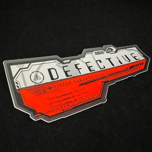 May include: A sticker with a futuristic design, featuring the word "DEFECTIVE" in bold white letters. The sticker has a red and gray color scheme with additional text including "TEST SUBJECT 004" and "GRAYSTAR INNOVATIONS".