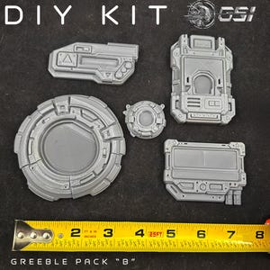 Könnte beinhalten: Ein graues DIY-Kit mit verschiedenen geometrischen Formen, darunter ein kreisförmiges Teil, rechteckige Paneele und andere Komponenten. Das Kit ist mit "Greeble Pack 'B'" beschriftet und enthält ein Maßband.